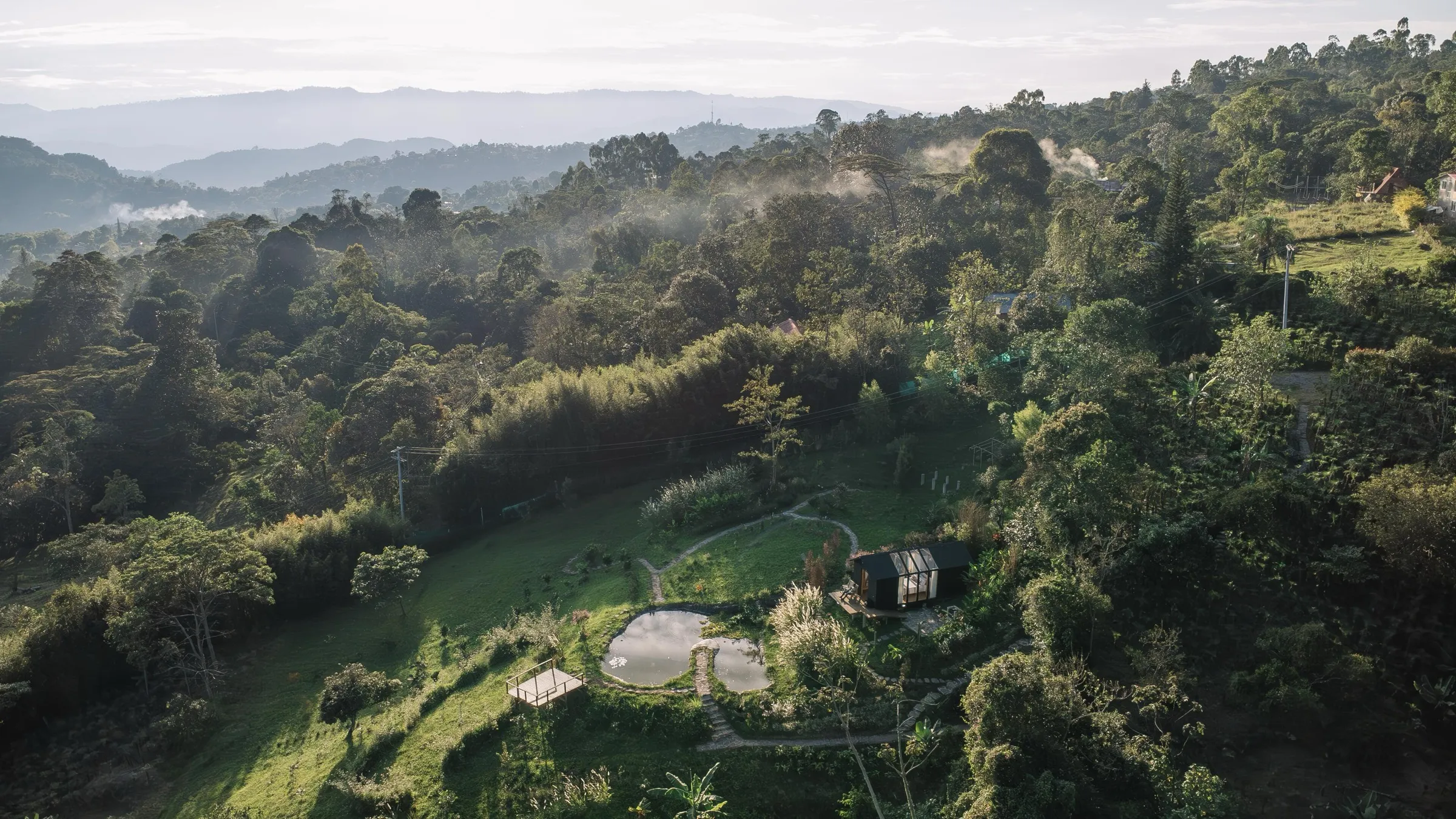 Vista aérea de una cabaña junto a un pequeño estanque entre montañas verdes y bosque, con neblina sobre el valle.