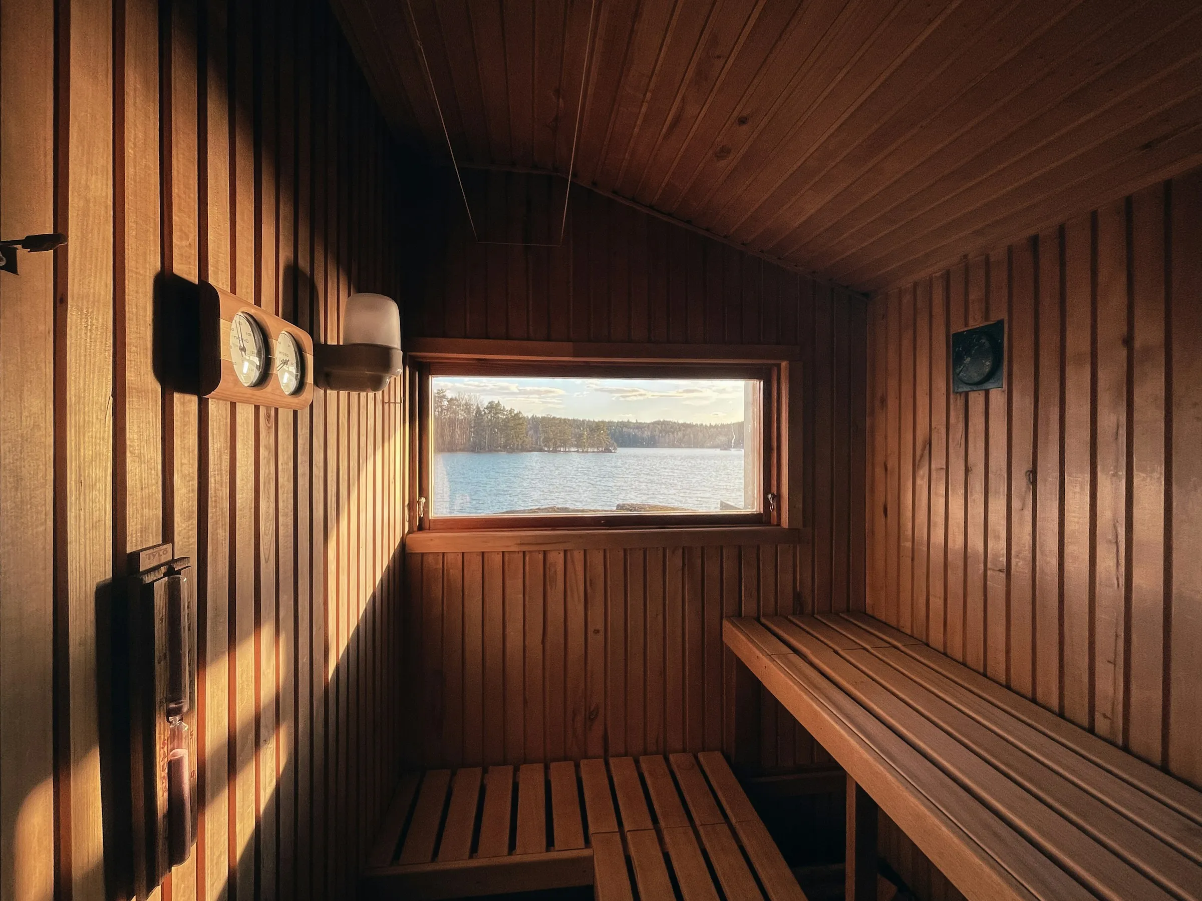 Interior de un sauna de madera con bancas y una ventana que deja ver un lago, iluminado por la luz cálida de la tarde.