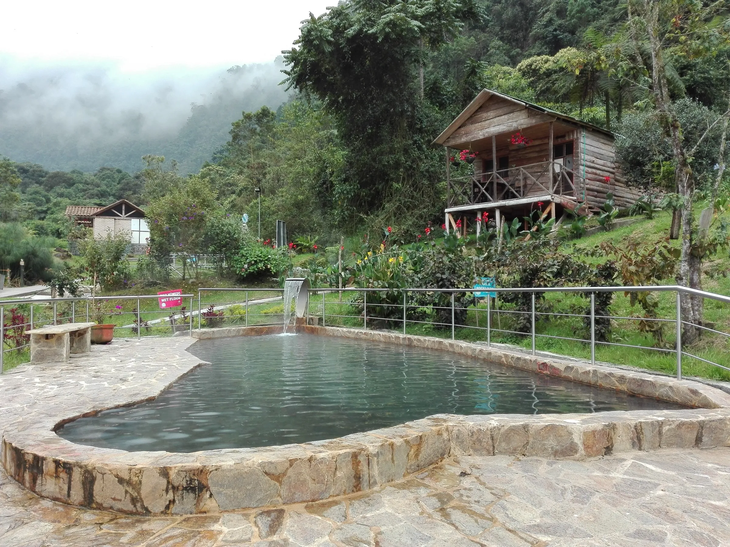 Piscina termal de borde de piedra rodeada de vegetación, con una cabaña de madera y montañas cubiertas de niebla al fondo.
